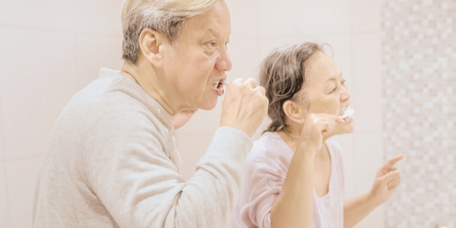 Duas pessoas idosas escovando os dentes. Parece que uma imita a outra ou ensina